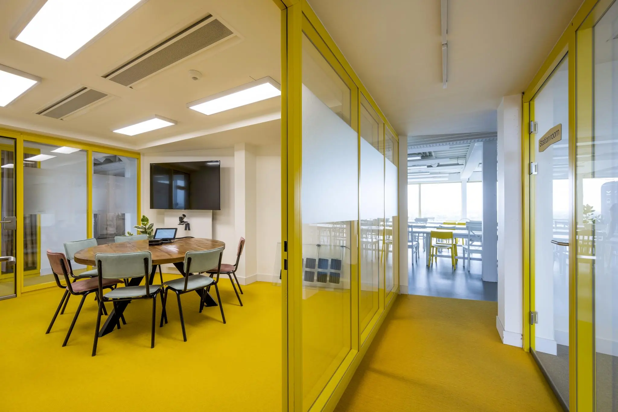 Moderne kantoorruimte aan de James Wattstraat met gele glazen wanden, een vergadertafel, stoelen en een flatscreen.